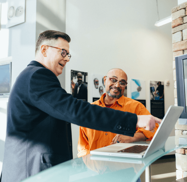 Two men stand by a glass desk with a laptop. One man, wearing a dark blazer, gestures at the laptop screen while the other, in an orange shirt, smiles and listens. Modern office setting with photographs on the wall.