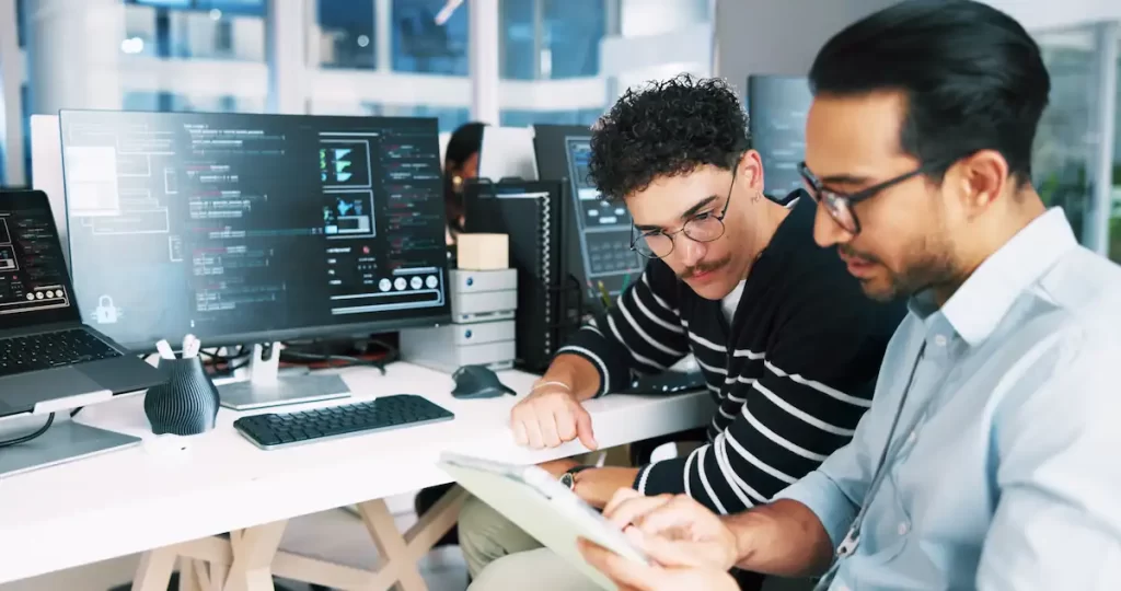 Two men sit together at a desk with computer monitors displaying code and data. One gestures at a tablet while the other looks on, highlighting collaborative problem-solving in a business office, possibly focused on IT support.