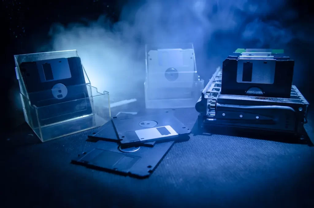 A collection of floppy disks is arranged on a dark surface, with some stacked in holders and others scattered in the foreground—an atmospheric nod to how today’s IT providers outgrow yesterday’s tech, all bathed in dramatic blue lighting and mist.