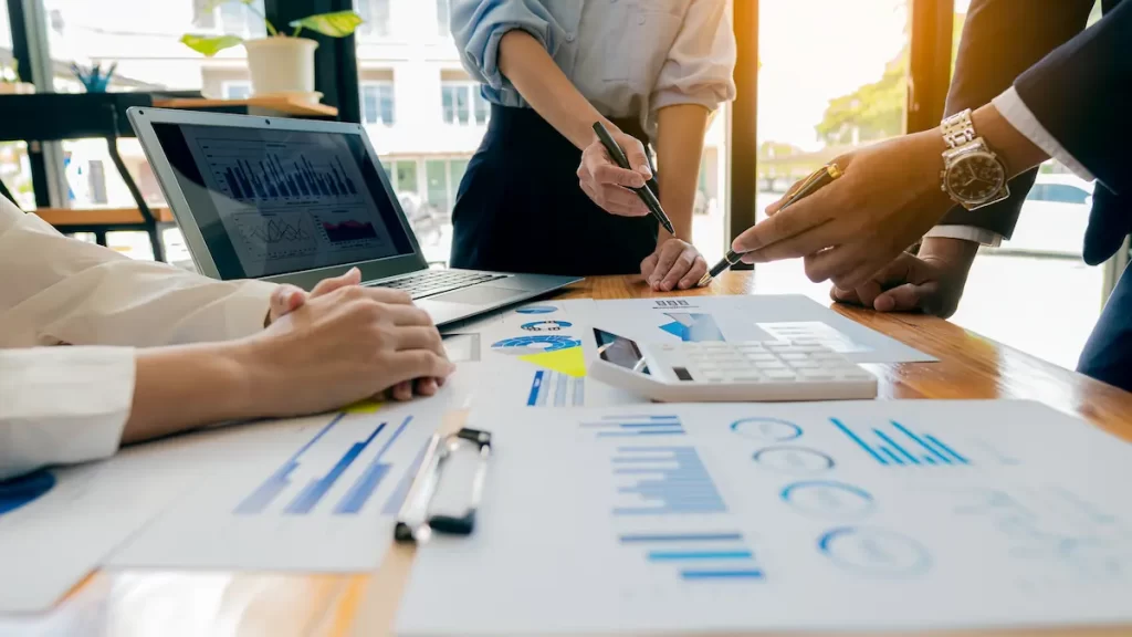 Three people collaborate at a table covered with business charts, graphs, a laptop, and a calculator, proactively discussing documents and IT solutions during a bright office meeting.