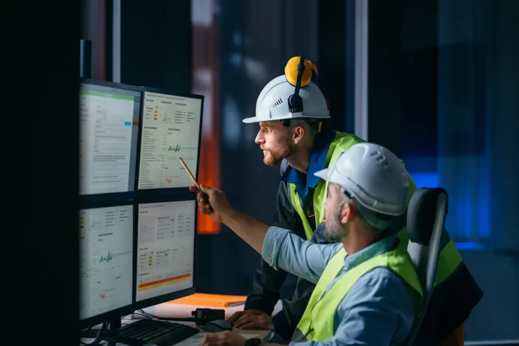Two engineers in safety vests and helmets review construction data and graphs on multiple computer monitors in a modern control room. One points to a screen with a pencil while the other observes attentively.