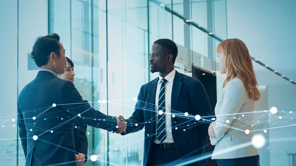 Four business professionals, two men and two women, stand in a modern Okanagan office. Two men are shaking hands while the women observe, as digital network lines overlay the image—symbolizing connection with an IT Partner or technology.