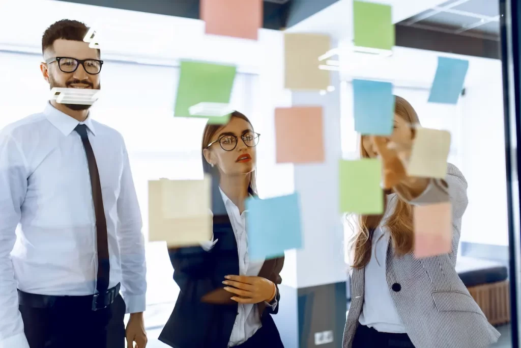 Three people in business attire stand by a glass wall covered with colorful sticky notes, discussing ideas. One person points to a note related to strategic IT planning while the others look on attentively.