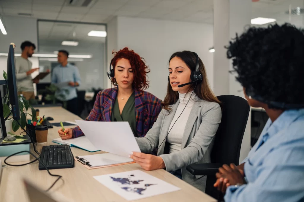 Three women wearing headsets sit at a desk in an office, looking at a document together, demonstrating how technology in the workplace can enhance collaboration. Two men are talking in the background. A computer, papers, and a plant are on the desk.