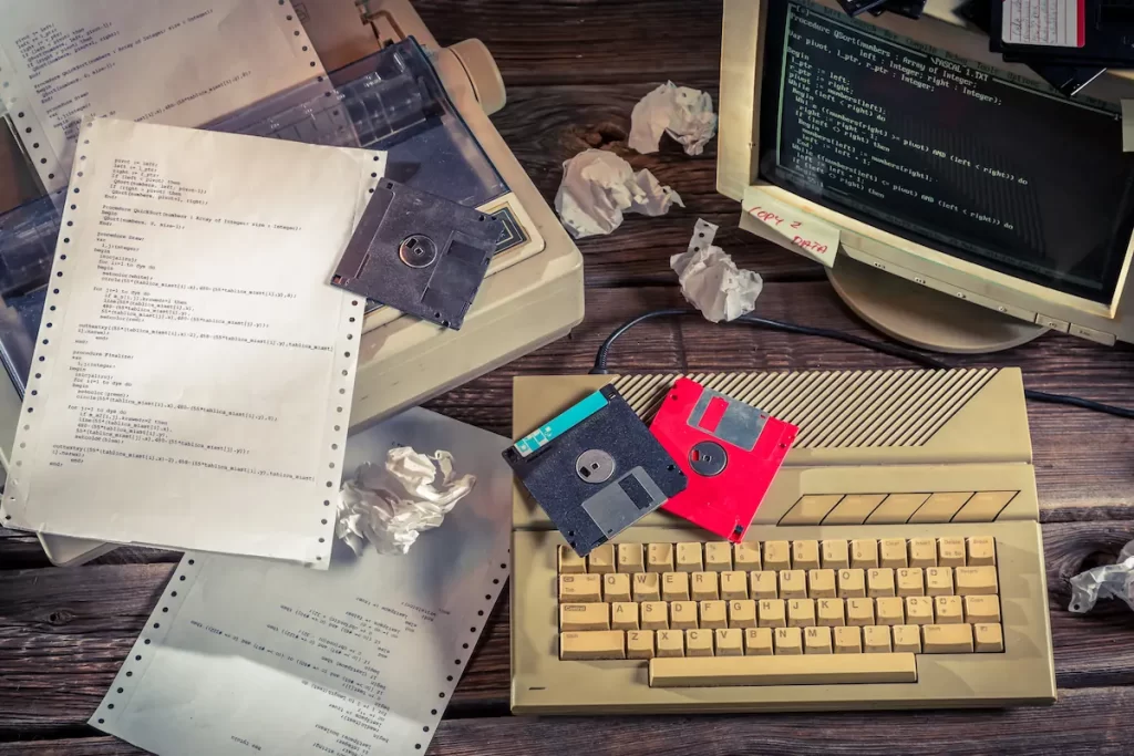 A vintage computer setup with a yellow keyboard, monitor displaying code, floppy disks, dot matrix printer paper, and several crumpled sheets on a wooden desk subtly evokes the era when technology debt began to accumulate—reminding us that legacy systems were once cutting-edge innovations.