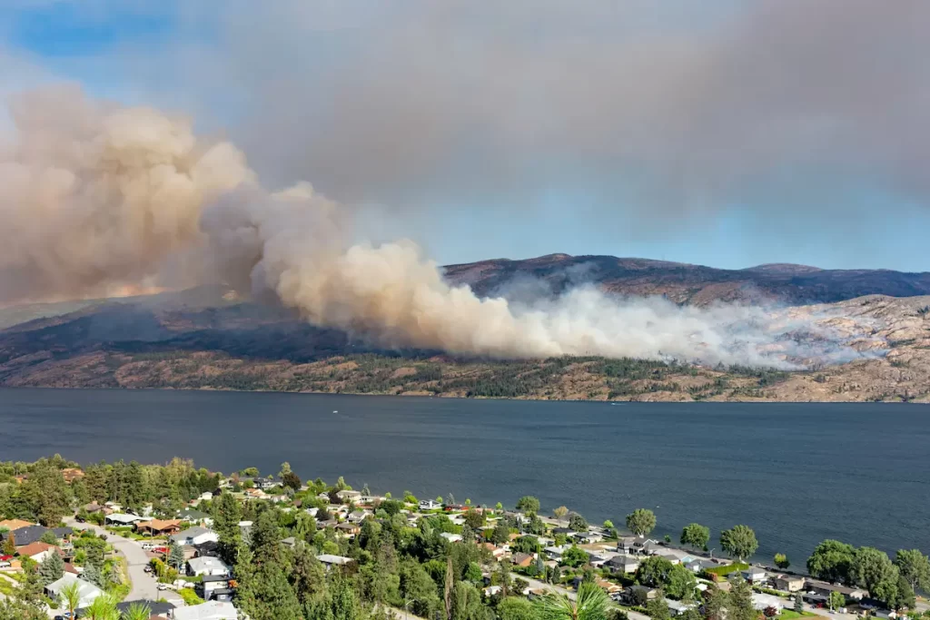 Smoke billows from a wildfire burning on a wooded hillside across a large body of water, with residential houses and trees in the foreground—a stark reminder of wildfire season in the Okanagan.