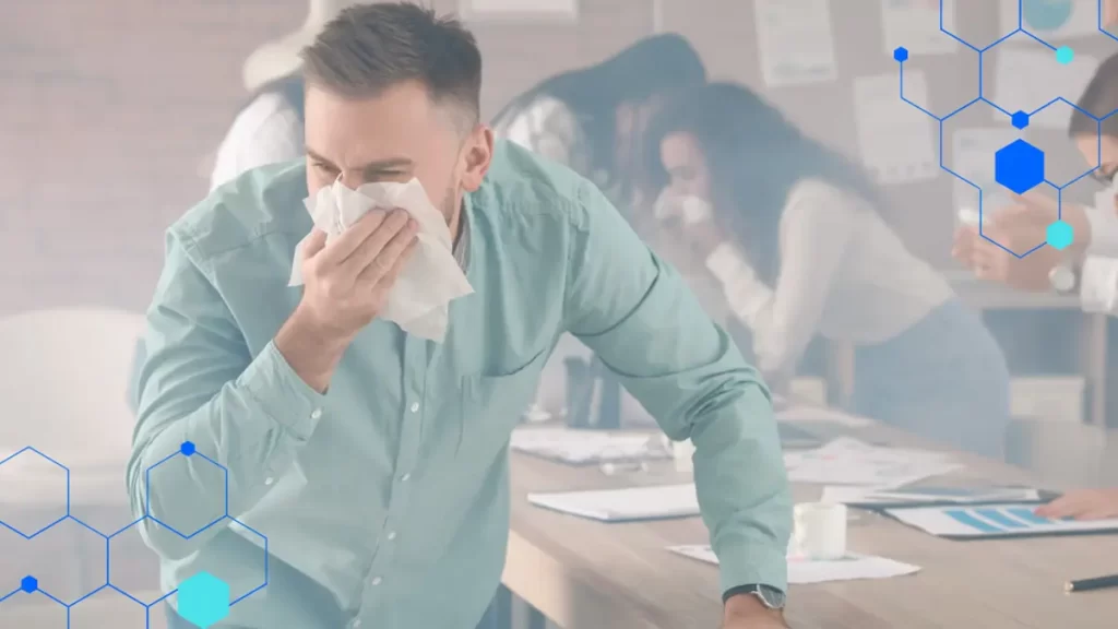 A man in a green shirt covers his nose and mouth with a tissue, appearing to sneeze—possibly due to wildfire smoke—while others in the background also look unwell in an office setting. Blue hexagon graphics overlay the image.